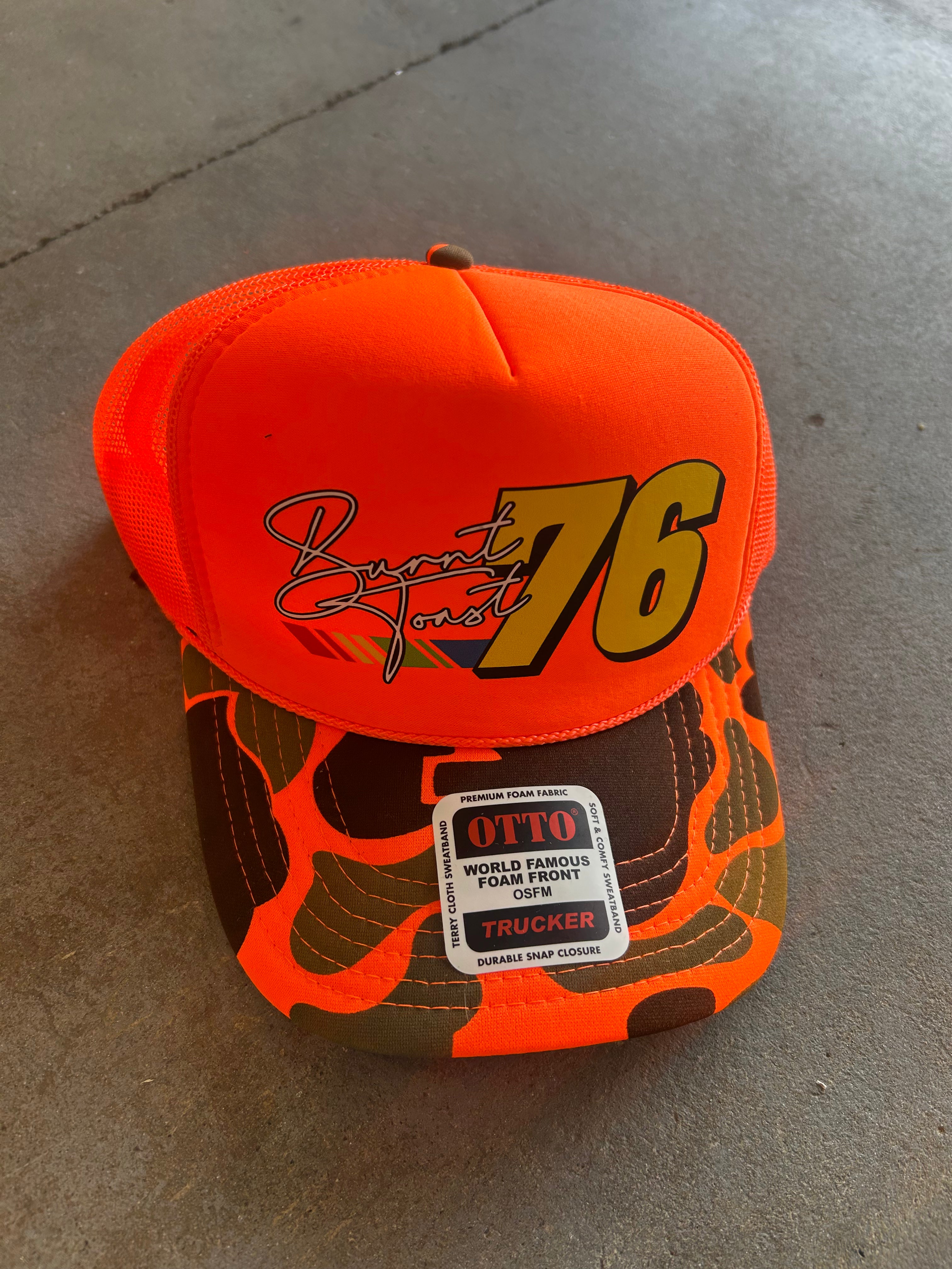 Orange trucker hat with camo brim and Burnt Toast 76 logo on the front, frontal view on a concrete surface