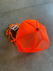 A bright neon orange trucker-style cap with a mesh back and a patterned front panel, resting on a gray concrete floor