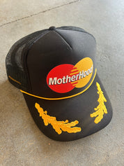 Black trucker hat with MotherHood logo and yellow detail, flat lay frontal view on a concrete surface
