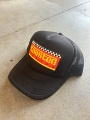 A black trucker hat with a curved brim, featuring a front patch that reads 'coasted racing' in yellow over a red background with a checkered flag stripe. The hat is photographed on a concrete floor