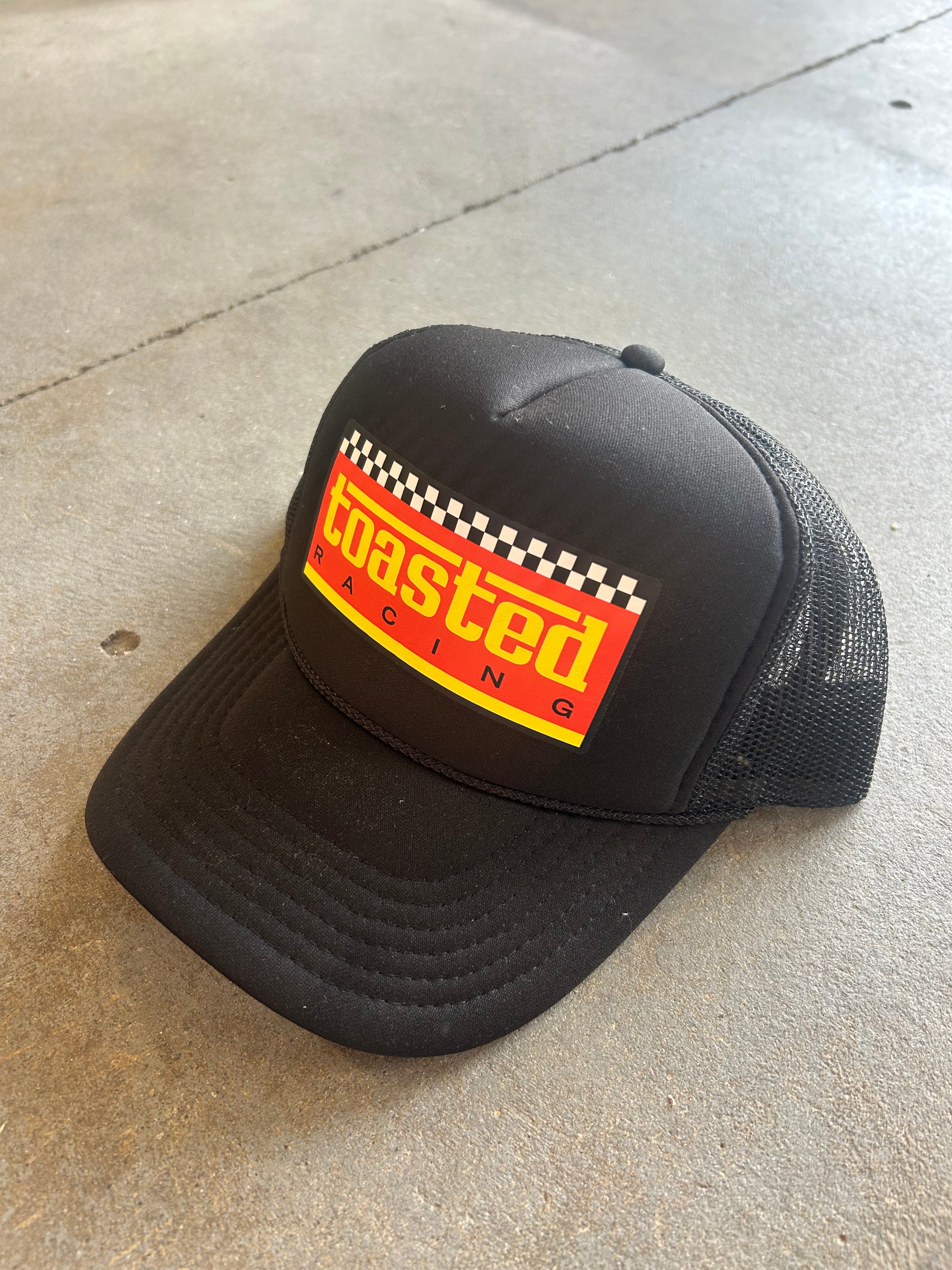 A black trucker hat with a curved brim, featuring a front patch that reads 'coasted racing' in yellow over a red background with a checkered flag stripe. The hat is photographed on a concrete floor