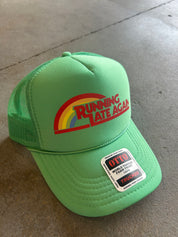 A bright green trucker hat with a rainbow graphic and the words 'RUNNING LATE AGAIN' on the front, resting on a gray concrete floor. A white and black OTTO sticker is visible on the brim.