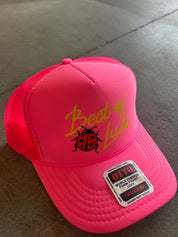 A bright pink trucker hat with yellow embroidered text reading 'Best of Luck' and a black ladybug graphic on the front. A white Otto brand sticker is attached to the brim, and the hat is photographed on a gray concrete floor