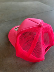 A bright neon pink trucker hat with a solid pink brim and pink mesh back panels, photographed on a gray concrete floor
