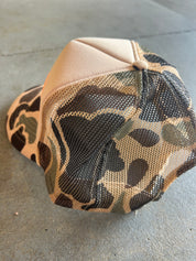 A baseball cap with a beige crown and a camouflage-patterned mesh back, resting on a gray concrete surface