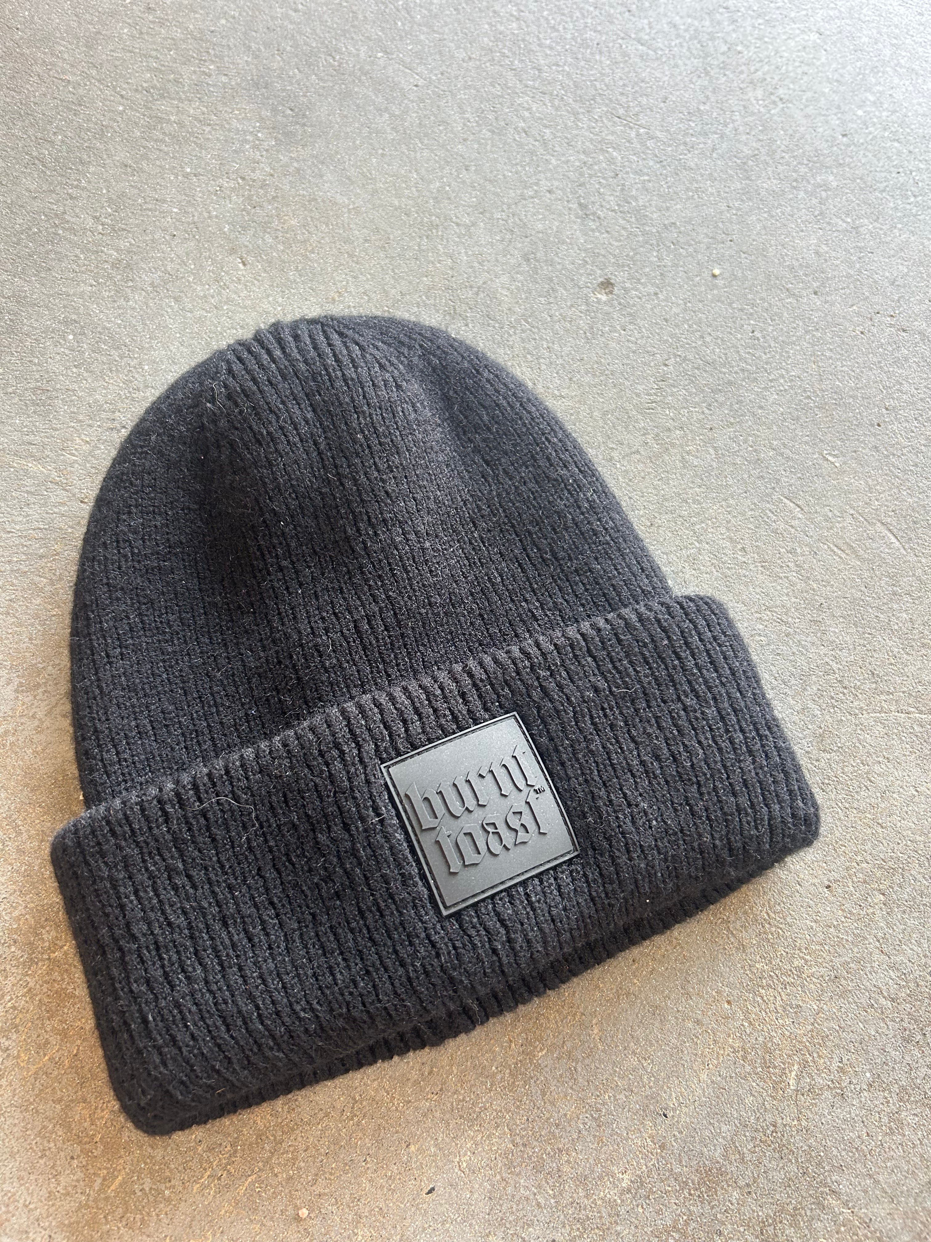 Black beanie with a Burnt Toast rubber logo on the brim on a concrete surface