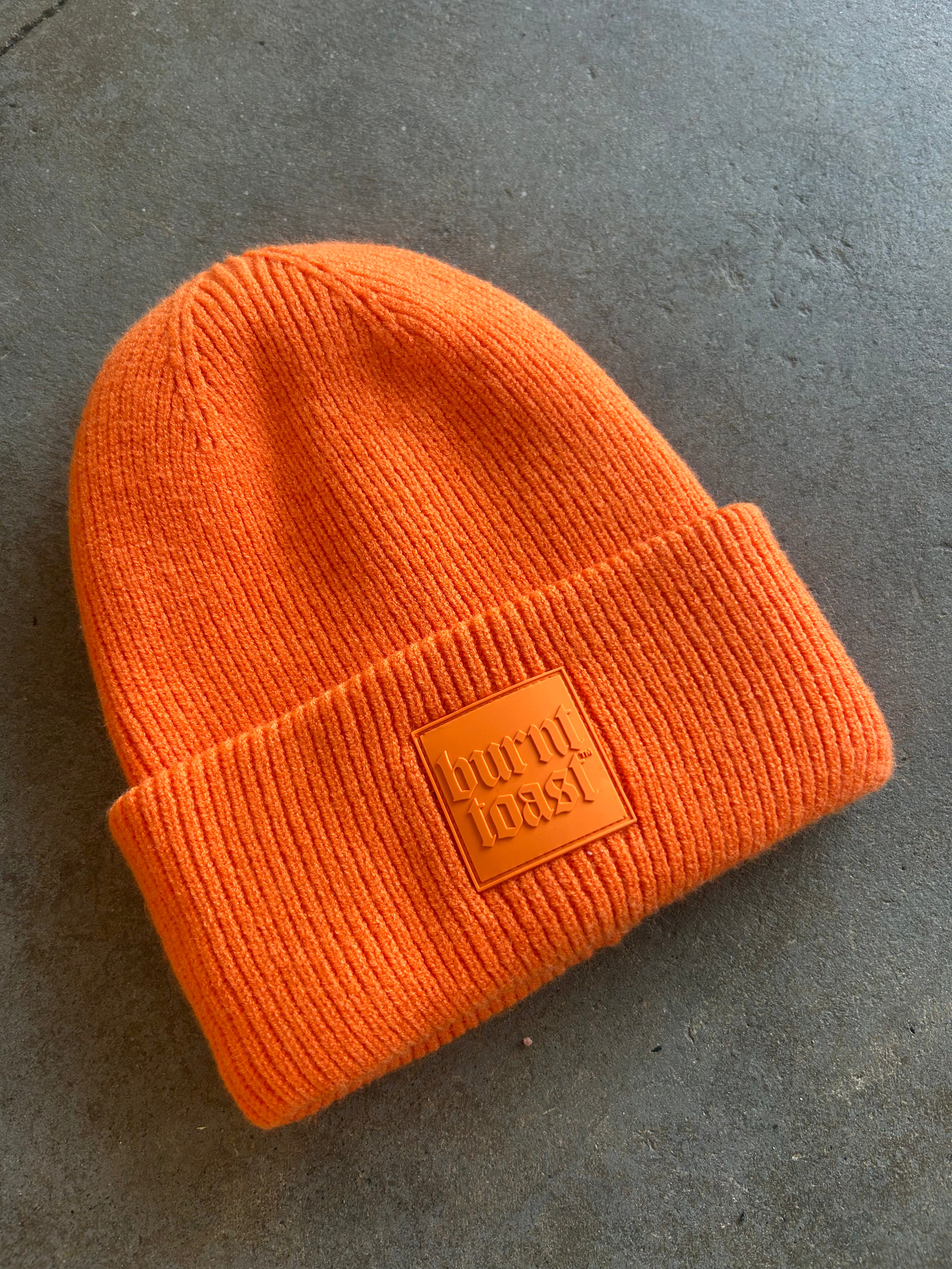 Orange beanie with a Burnt Toast rubber logo on the brim on a concrete surfaceO