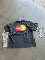 A black long-sleeve t-shirt with a 'Motherhood' parody logo designed to look like the Mastercard logo, laid flat on a concrete floor. The sleeves feature a camouflage print