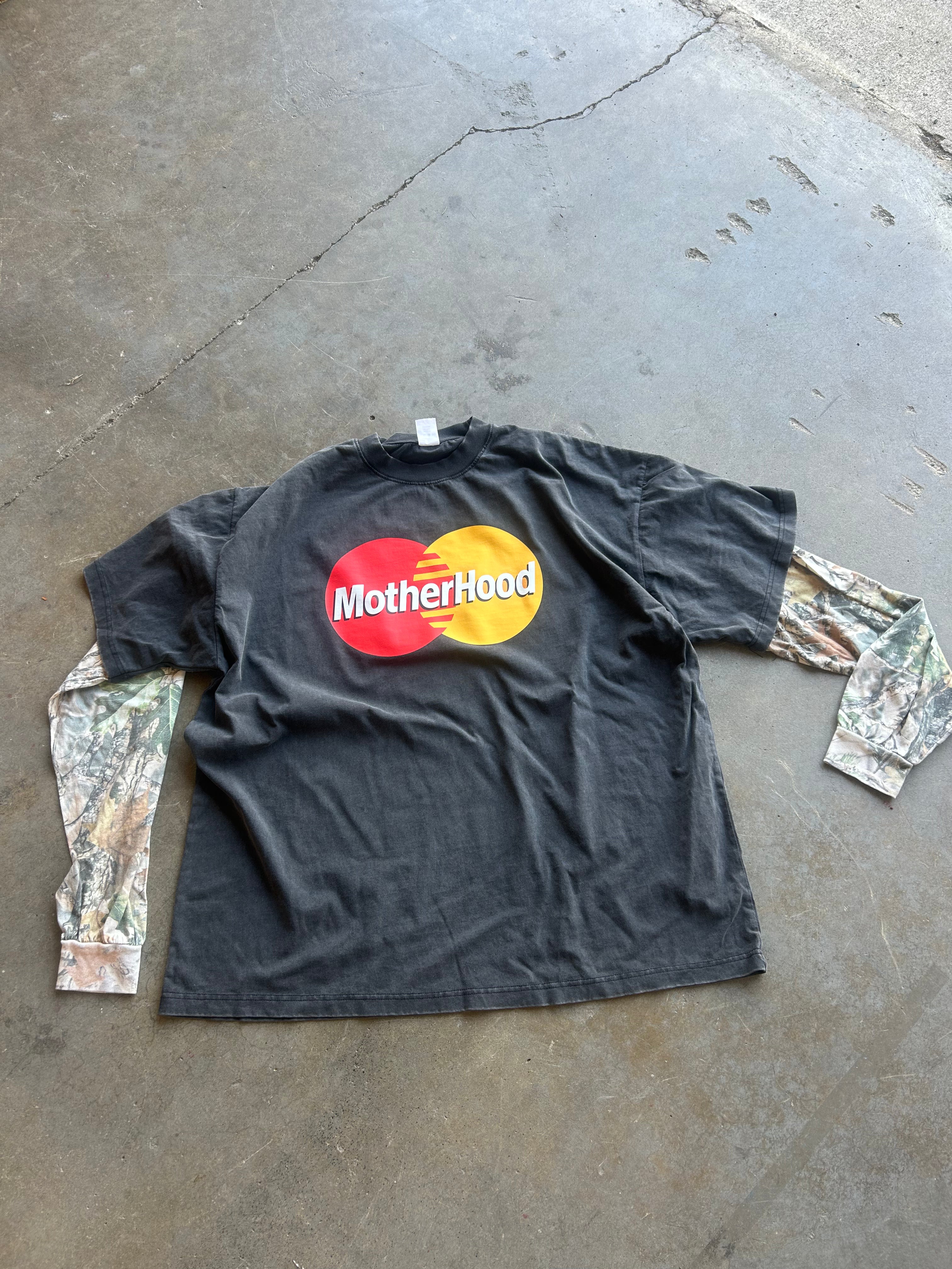 A black long-sleeve t-shirt with a 'Motherhood' parody logo designed to look like the Mastercard logo, laid flat on a concrete floor. The sleeves feature a camouflage print