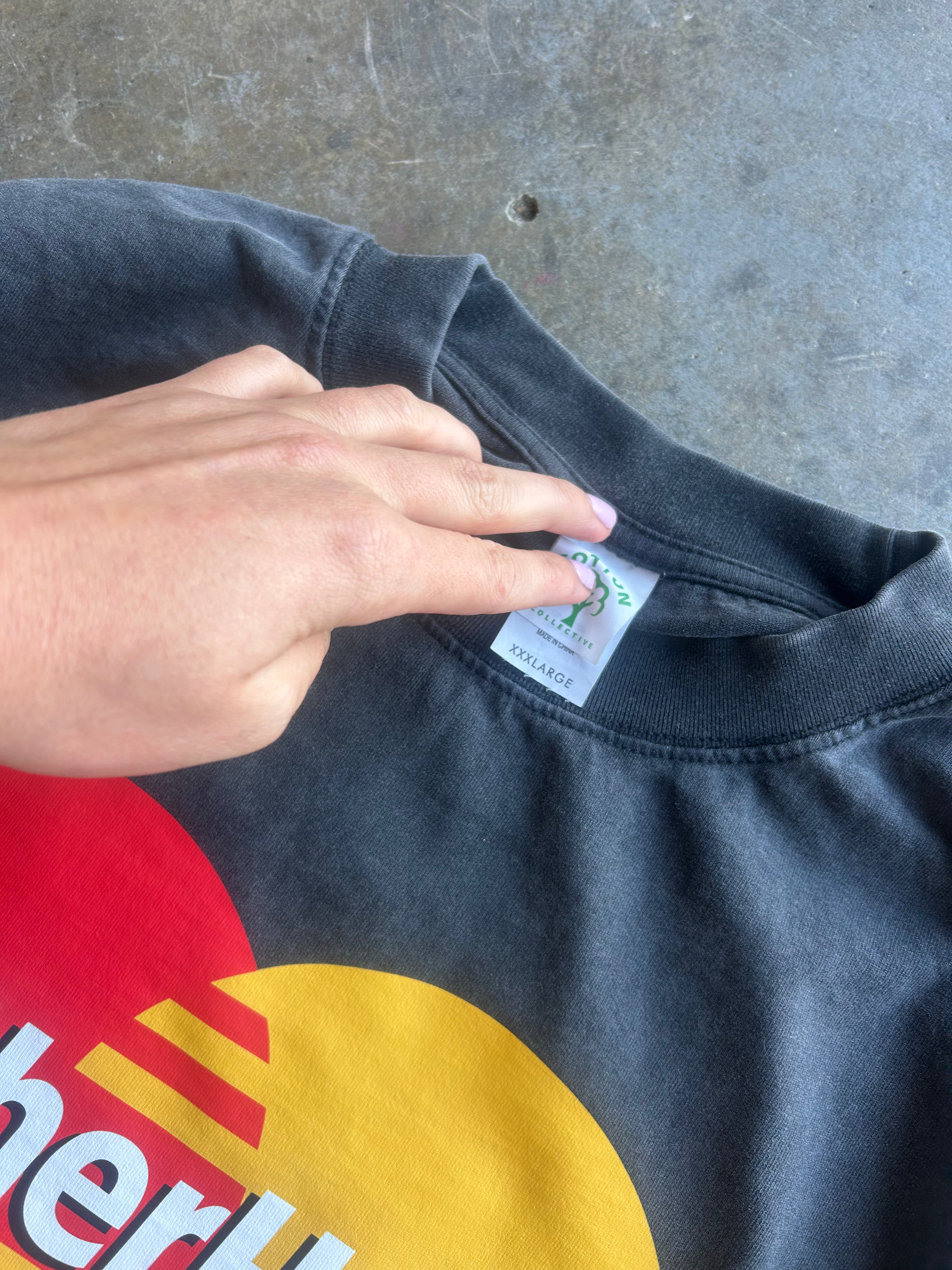 A close-up of a person's hand pointing to a white brand tag on the collar of a faded black t-shirt. The shirt features a large red and yellow circular graphic on the front with white text partially visible