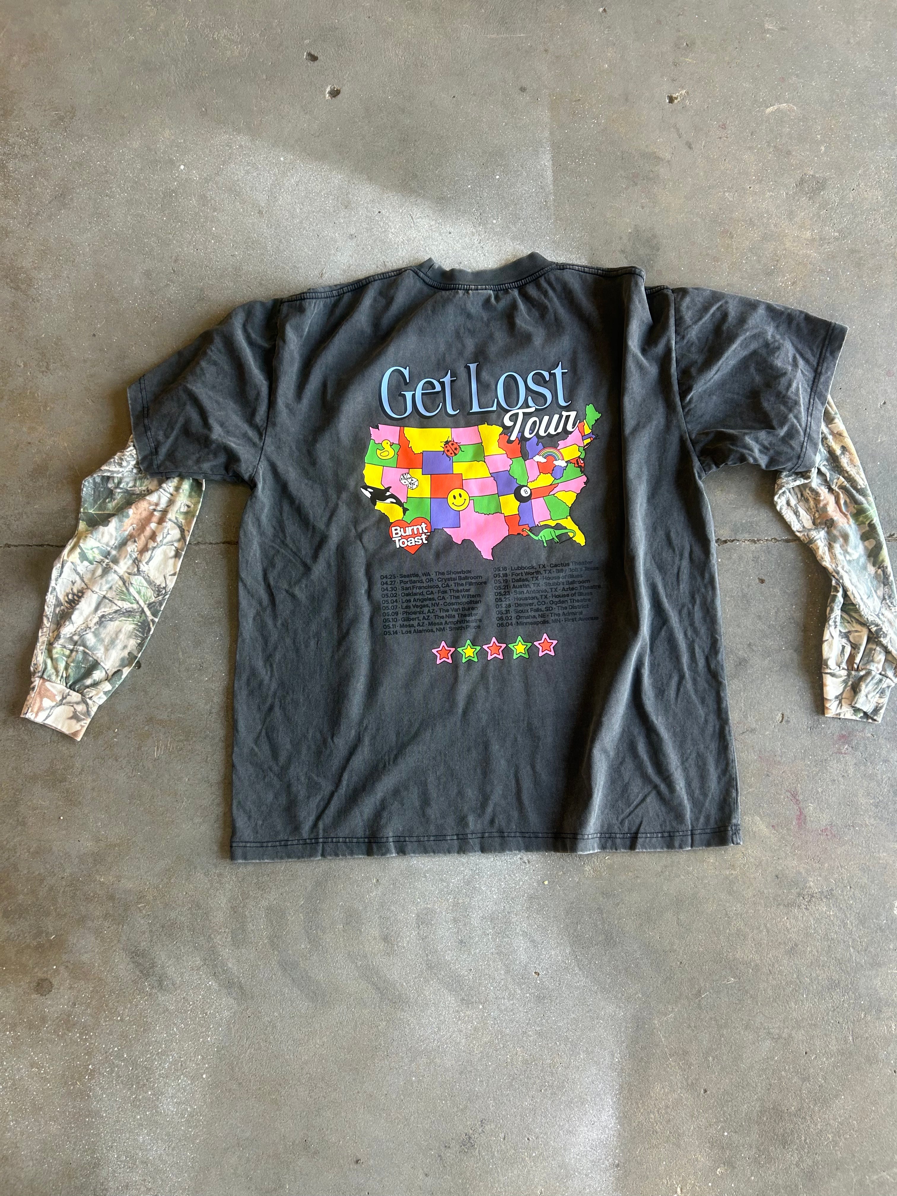 A black graphic t-shirt laid flat on a concrete floor, featuring a colorful 'Get Lost Fun' USA map design on the back with camouflage-print long sleeves