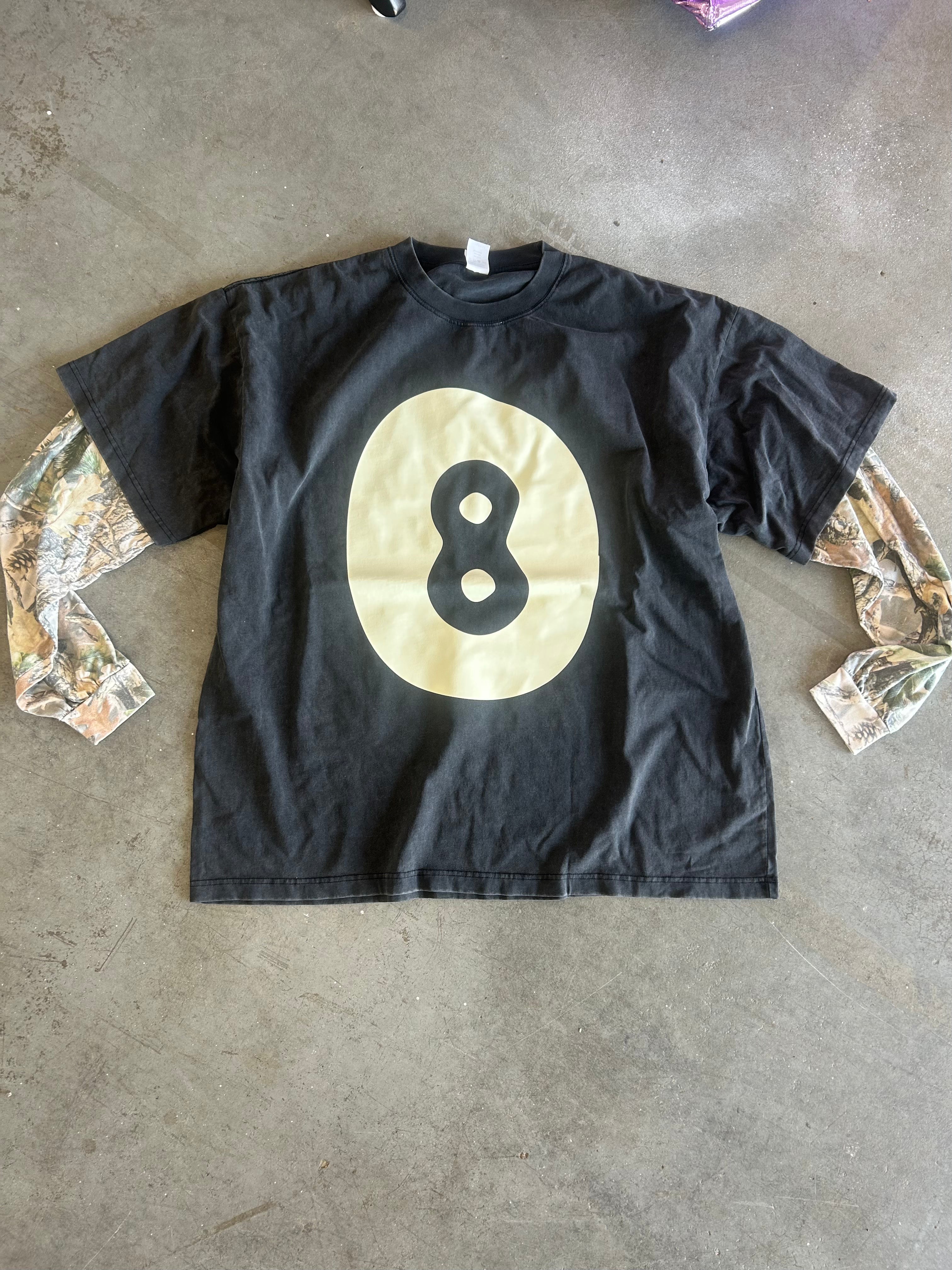 Long sleeve grey shirt with camo sleeves and creme 8 ball logo in the middle, on a concrete surface