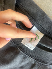 A close-up of a person's hand pointing to a white size label on the inside collar of a dark gray sweatshirt, showing the brand 'Cotton Collective' and size 'XXL LARGE'