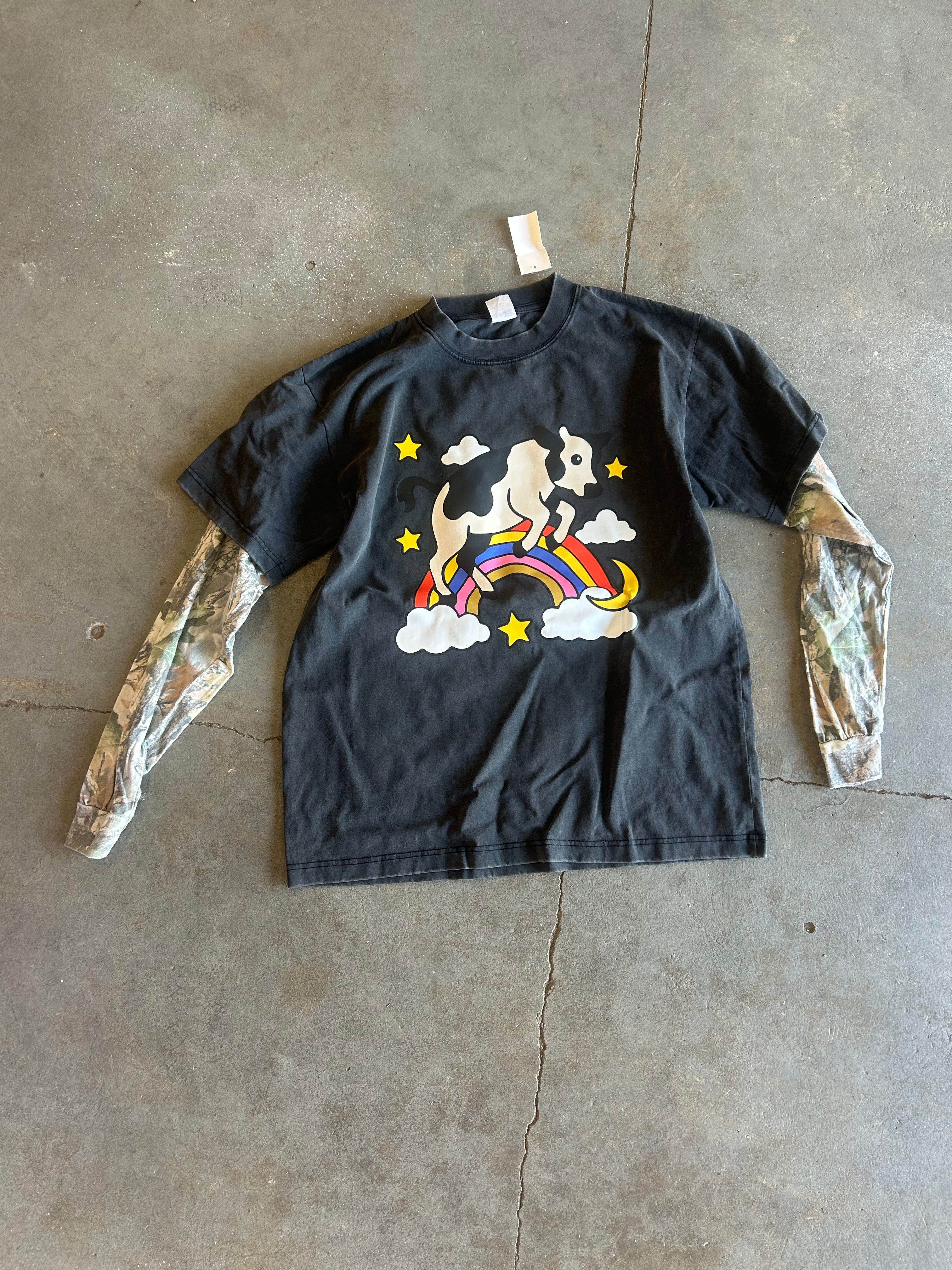 A black short-sleeve t-shirt with a graphic of a dog riding a rainbow on clouds, styled with camouflage long sleeves, laid flat on a concrete floor