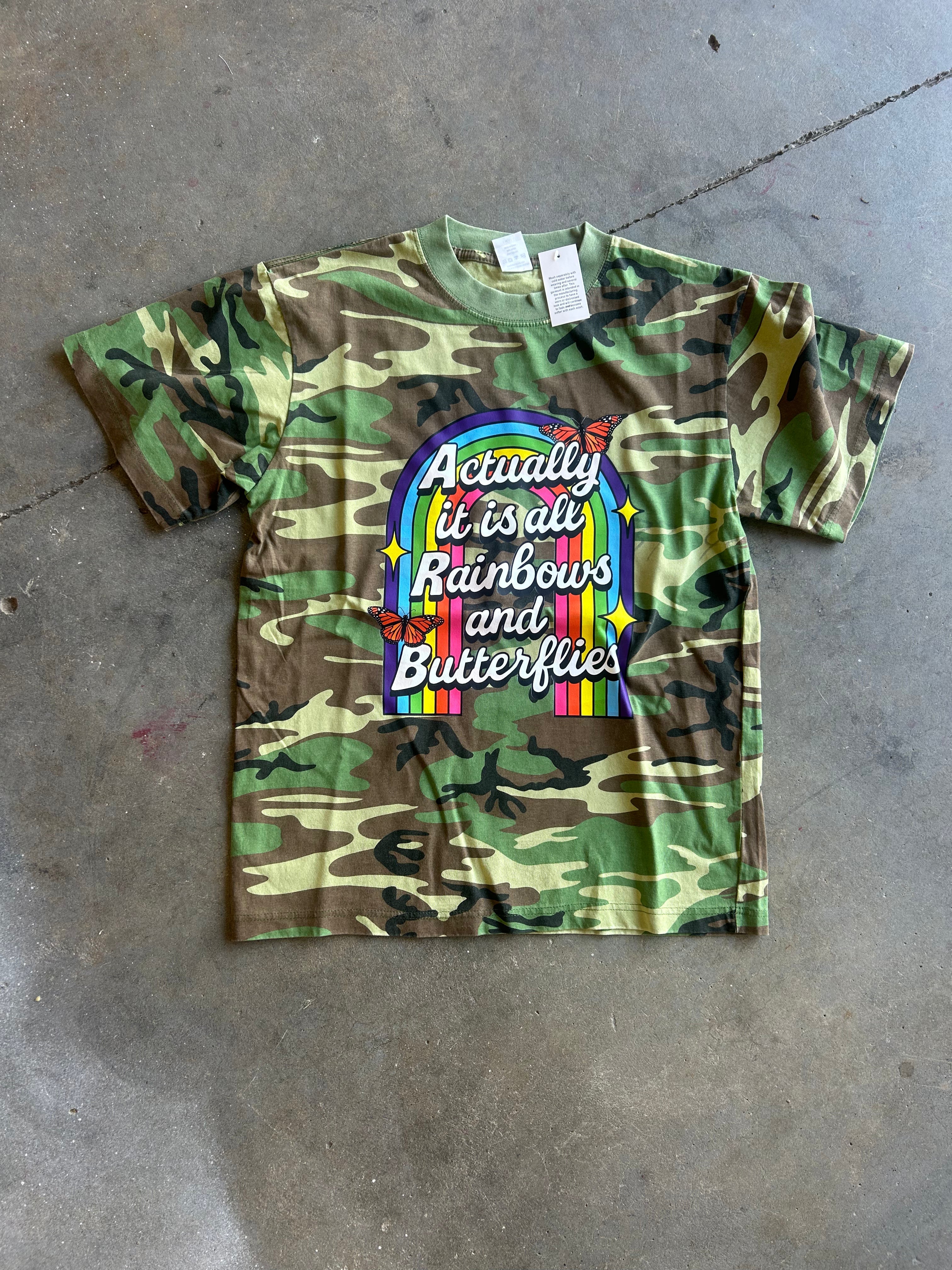 A short-sleeve kids' t-shirt in a green and brown camouflage pattern, featuring a colorful graphic on the front that reads 'Actually it is all Rainbows and Butterflies' inside a rainbow arch. The shirt is laid flat on a gray concrete floor
