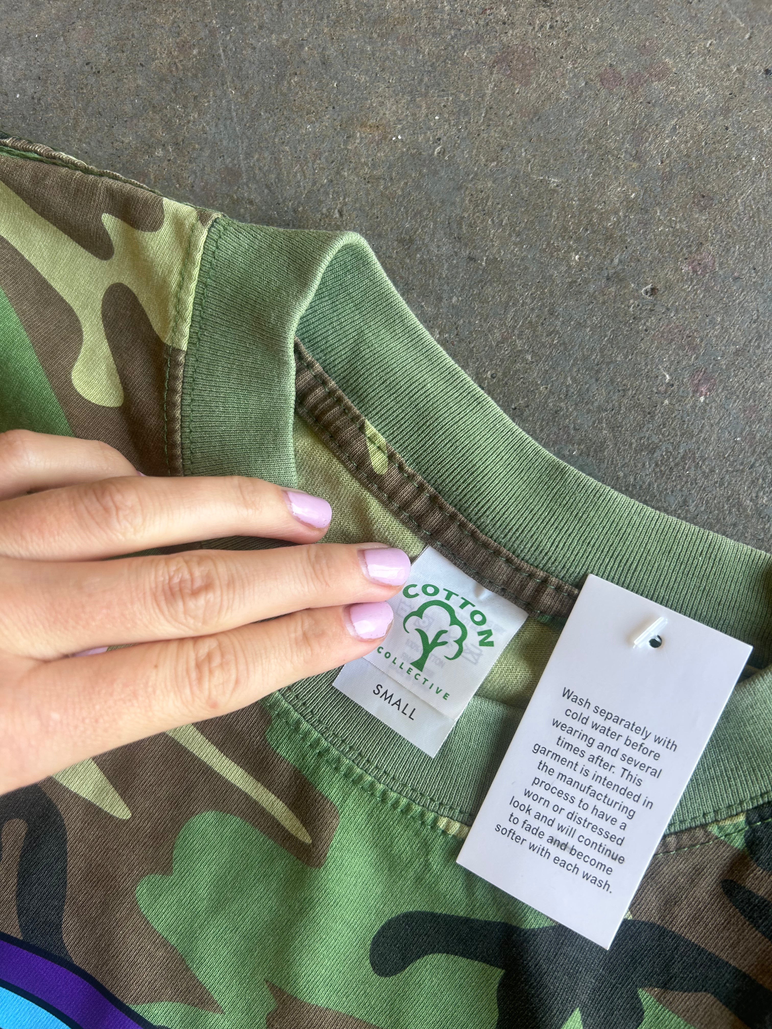 Close-up of a camouflage crewneck sweatshirt with a hand pointing to the neck label, which reads Cotton Star, on a concrete floor.