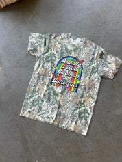 A short-sleeve camouflage t-shirt laid flat on a gray concrete floor. The shirt features a colorful graphic in the center with a rainbow arch and the text 'Actually It is all Rainbows and Butterflies' in white script.