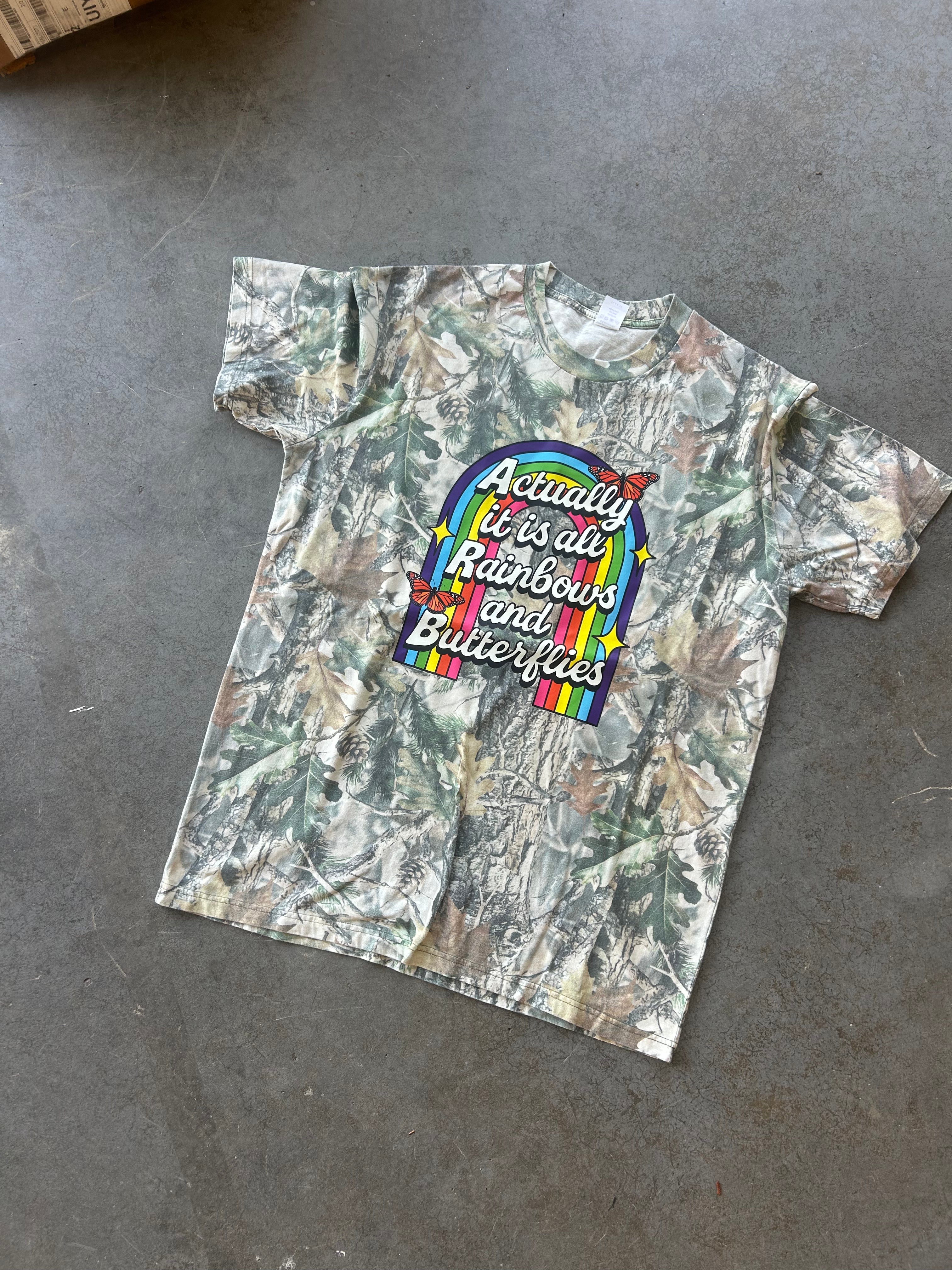 A short-sleeve camouflage t-shirt laid flat on a gray concrete floor. The shirt features a colorful graphic in the center with a rainbow arch and the text 'Actually It is all Rainbows and Butterflies' in white script.