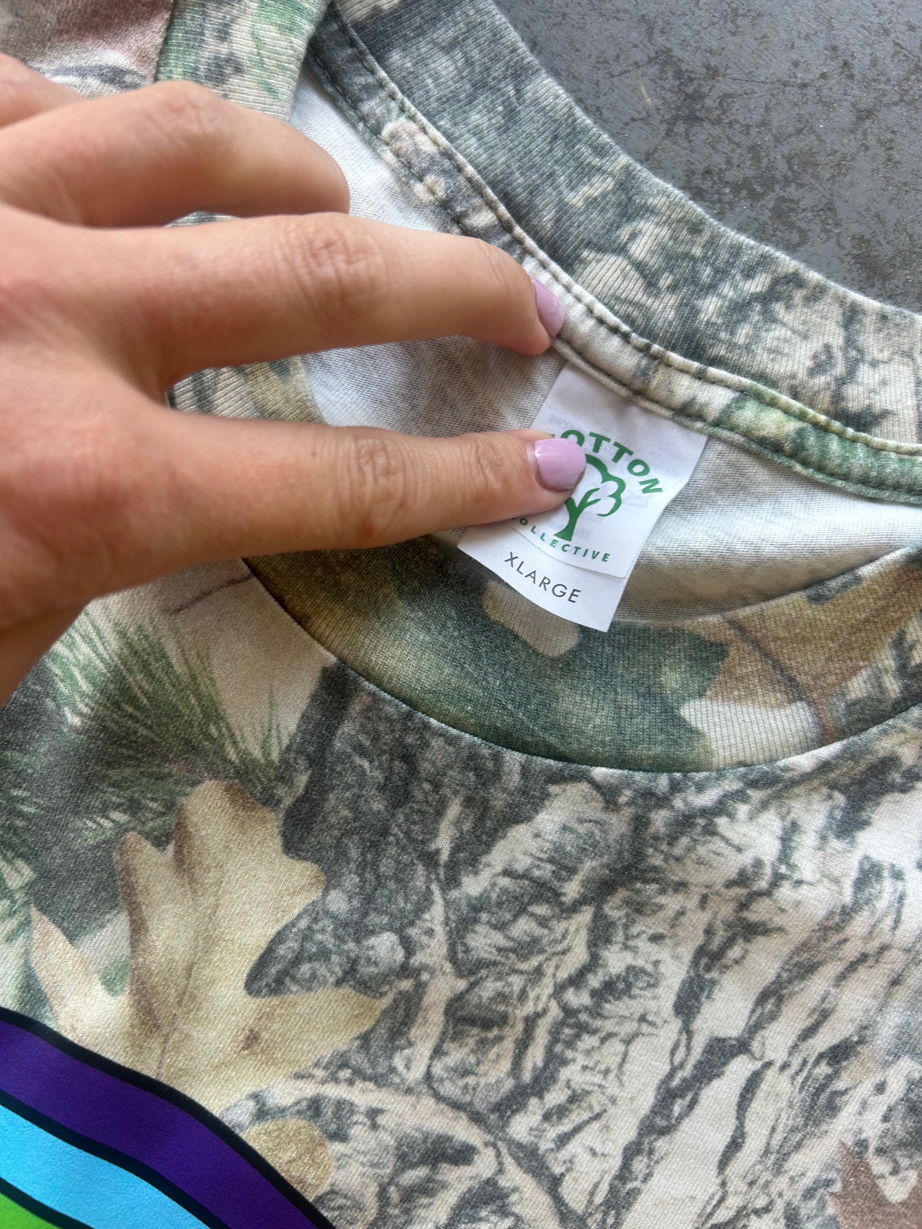 Close-up of a camouflage t-shirt with a hand pointing to the inner neck label which reads 'COTTON PRACTICE' and 'X-LARGE'.