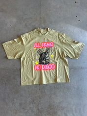 A light olive green graphic t-shirt laid flat on a gray concrete floor. The shirt features a central graphic of a black cat with yellow stars, framed by the text 'ALL PANIC NO DIBBO' in a hot pink bubble font.