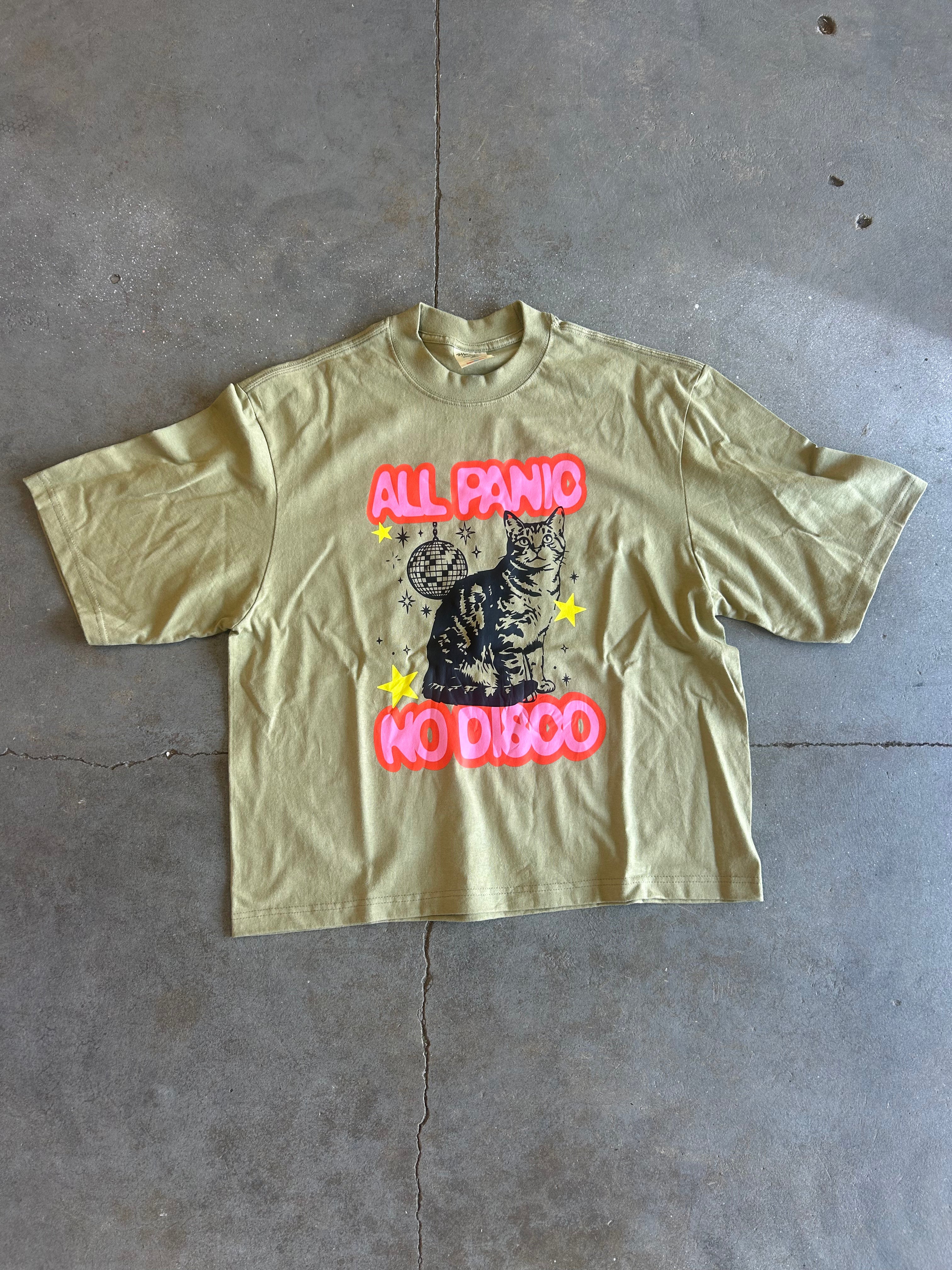 A light olive green graphic t-shirt laid flat on a gray concrete floor. The shirt features a central graphic of a black cat with yellow stars, framed by the text 'ALL PANIC NO DIBBO' in a hot pink bubble font.