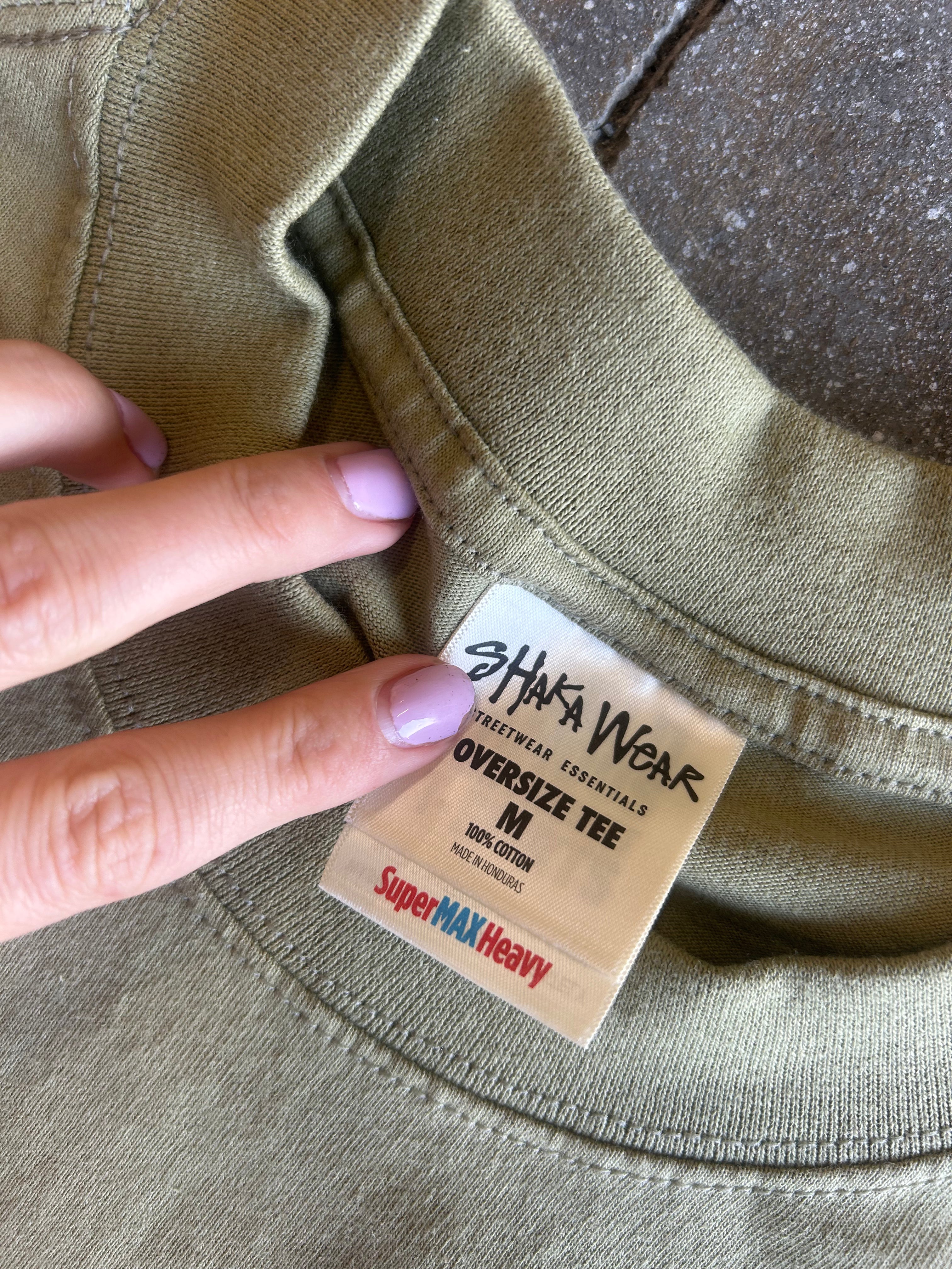 Close-up of an oversized olive green t-shirt neck label being held by a hand with lavender-painted fingernails. The label reads 'SHAKA WEAR' and 'Oversize Tee M'.