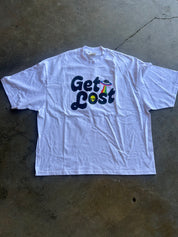 A white short-sleeve t-shirt with a graphic reading 'Get Lost' in dark blue bubble letters, featuring a small rainbow and yellow smiley face, laid flat on a concrete floor.