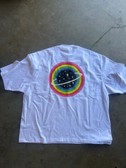A white short-sleeve t-shirt laid flat on a concrete floor, featuring a colorful graphic on the back with concentric rainbow rings, a black starry space circle, and a white ring-like orbit.
