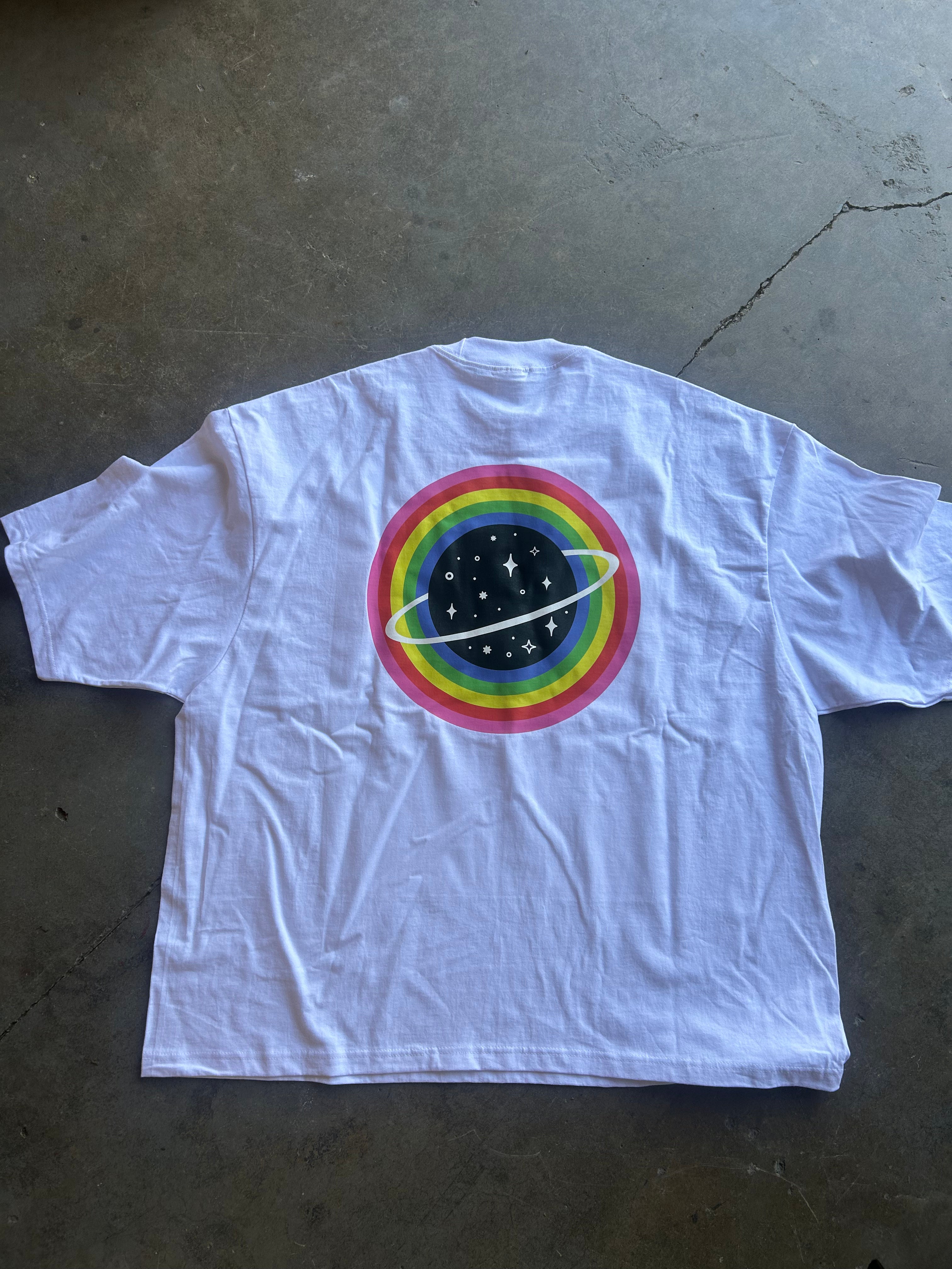 A white short-sleeve t-shirt laid flat on a concrete floor, featuring a colorful graphic on the back with concentric rainbow rings, a black starry space circle, and a white ring-like orbit.
