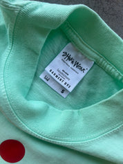 Close-up of a mint green garment dye shirt with a white inner neck label reading 'SHAKA WEAR' and 'GARMENT DYE', plus a red circular dye patch visible on the lower left.
