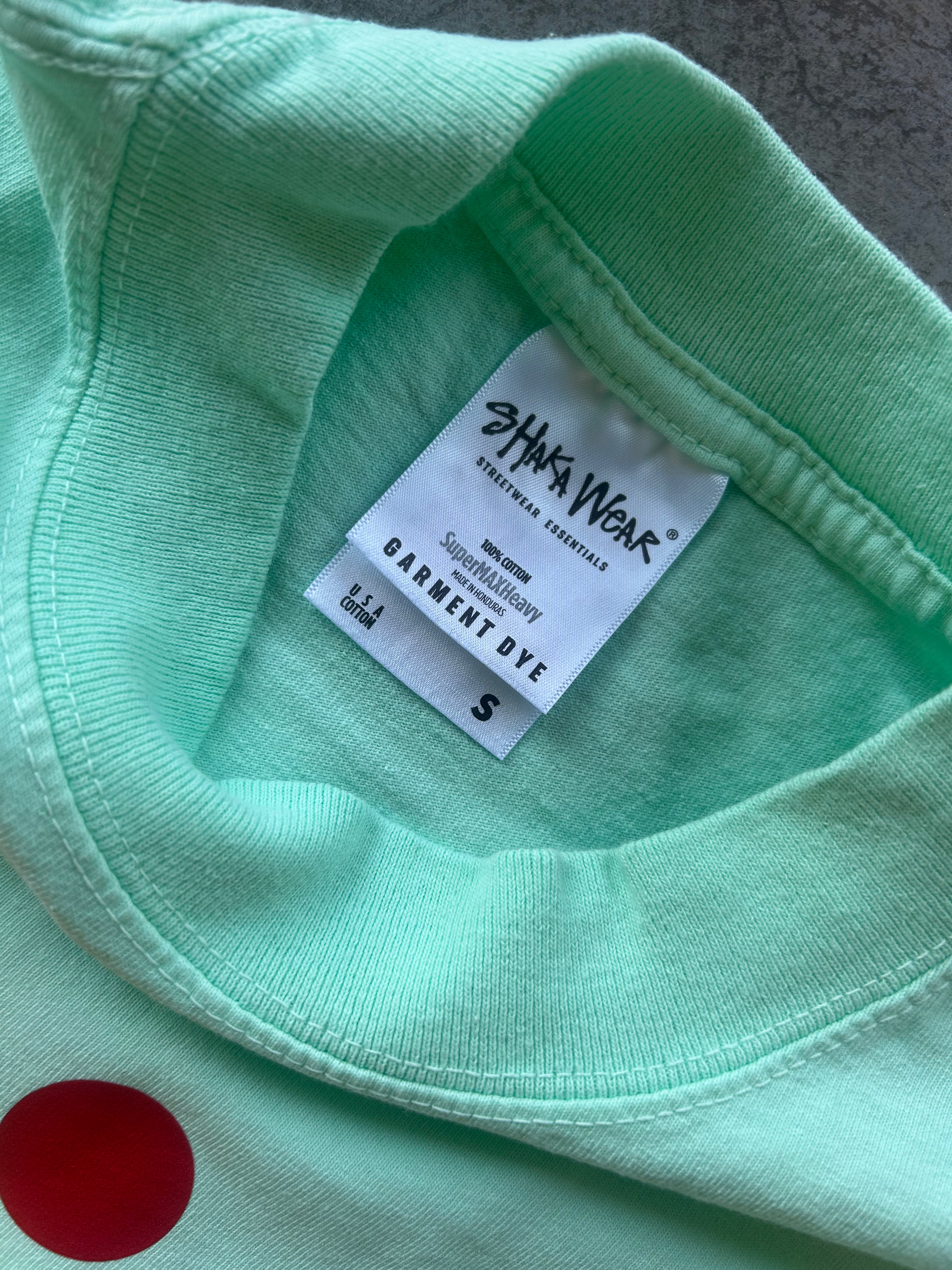 Close-up of a mint green garment dye shirt with a white inner neck label reading 'SHAKA WEAR' and 'GARMENT DYE', plus a red circular dye patch visible on the lower left.