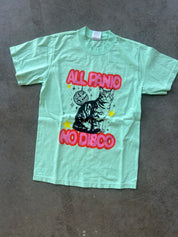A mint green graphic t-shirt laid flat on a gray concrete floor. The shirt features a black cat illustration sitting in front of a disco ball, with the text 'ALL PANIC NO DISCO' in red bubble letters and yellow stars.