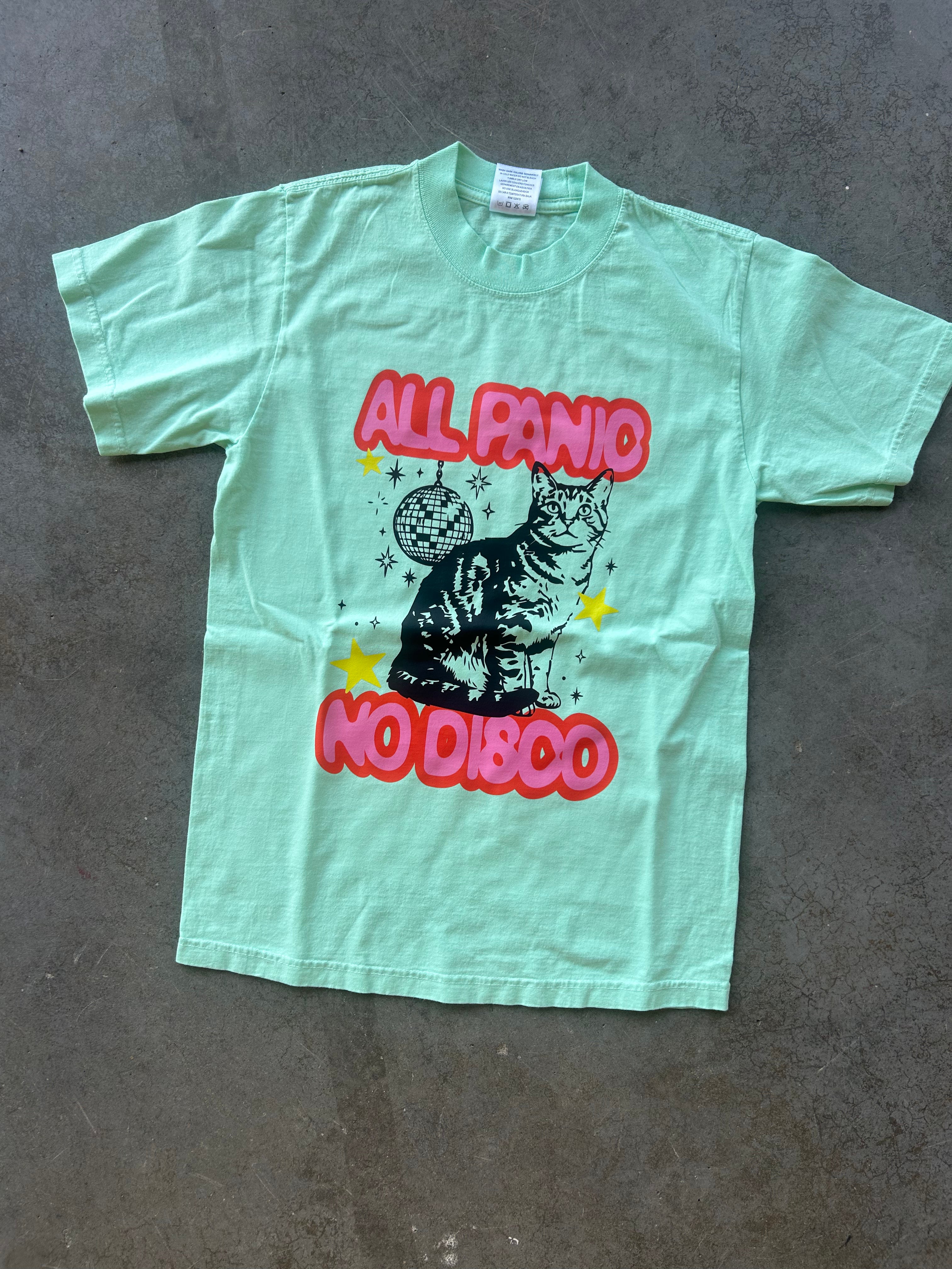 A mint green graphic t-shirt laid flat on a gray concrete floor. The shirt features a black cat illustration sitting in front of a disco ball, with the text 'ALL PANIC NO DISCO' in red bubble letters and yellow stars.