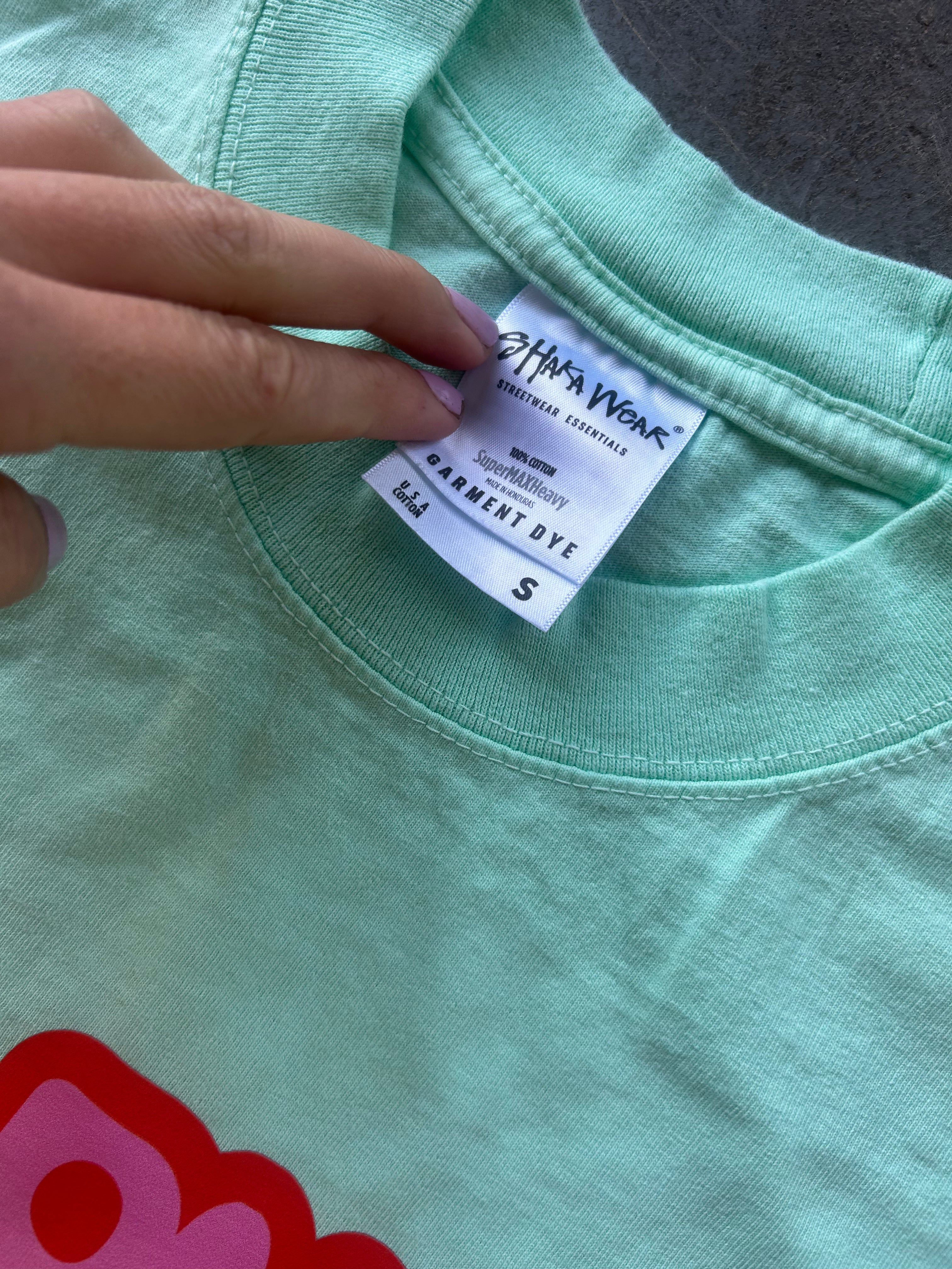 A close-up of a mint green t-shirt showing the inner neck label which reads 'Chic Wear' and 'Permanent Dye', with a hand pointing to the label.