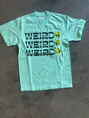 A mint green short-sleeve t-shirt laid flat on a concrete floor, featuring the word 'WEIRD' printed three times in black with yellow alien heads replacing the letters.