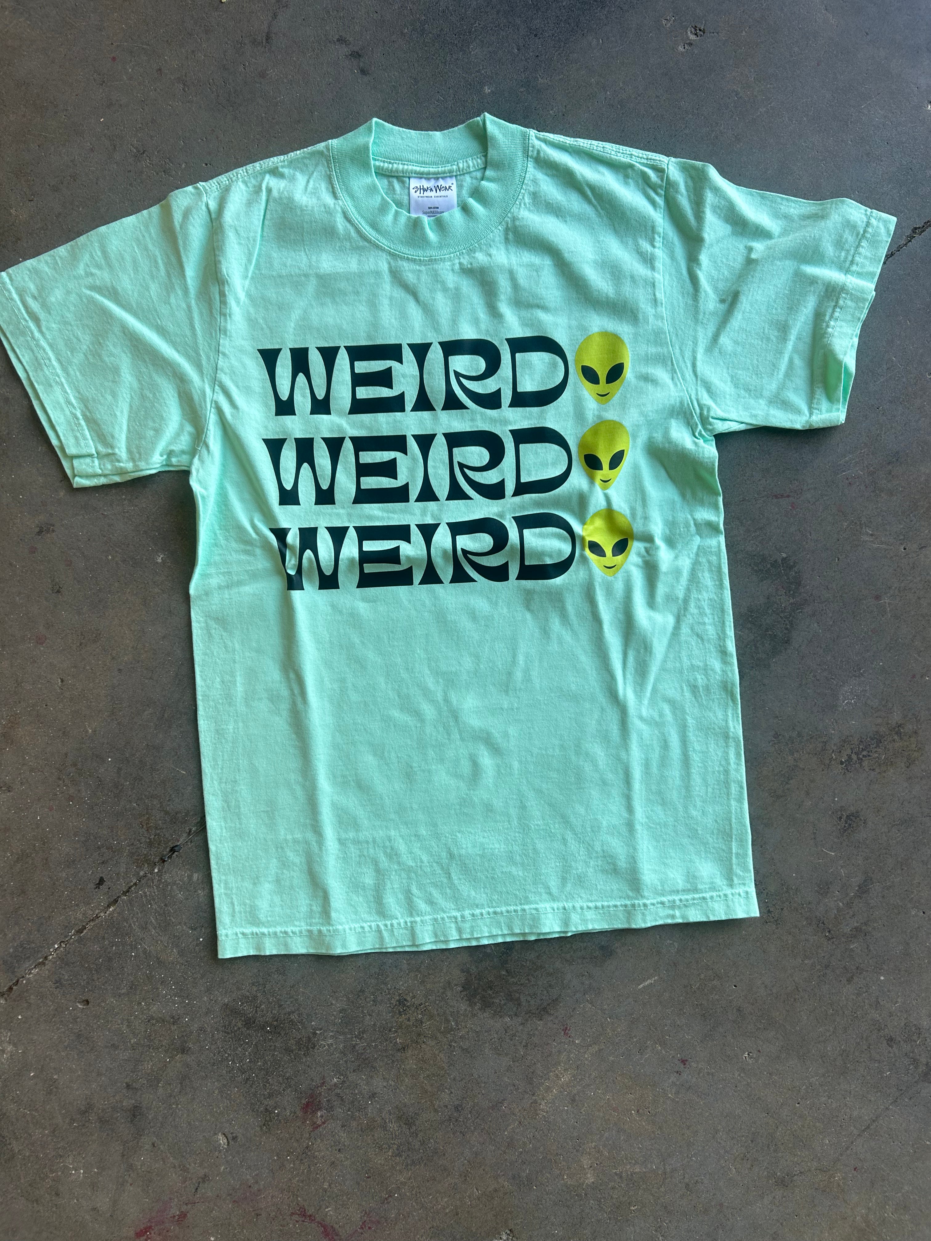 A mint green short-sleeve t-shirt laid flat on a concrete floor, featuring the word 'WEIRD' printed three times in black with yellow alien heads replacing the letters.