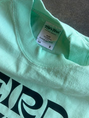 Close-up of a mint green t-shirt with a black graphic print, showing the inner neck label for 'SMA Wear' and size 'S'.