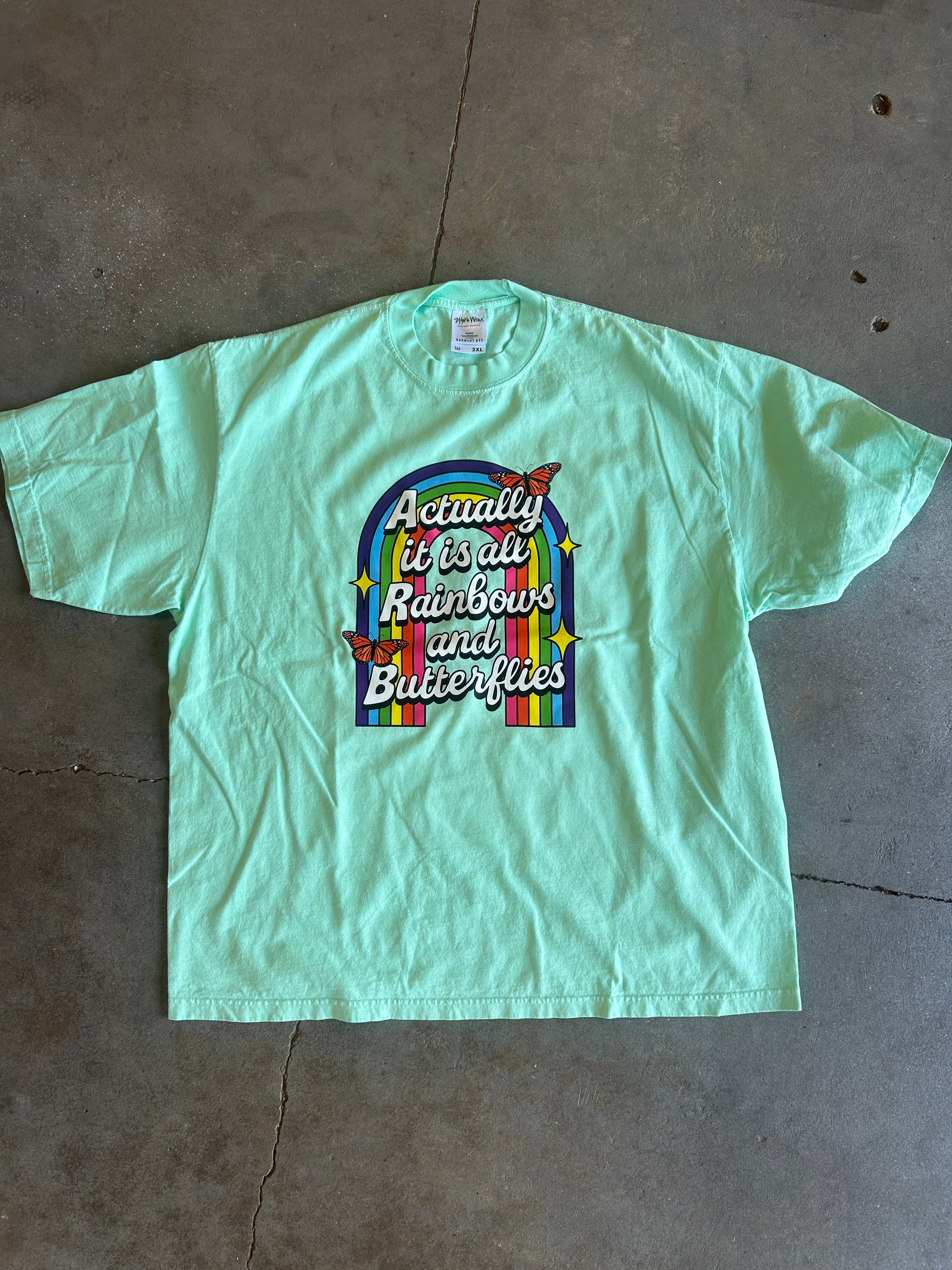 A mint green short-sleeve t-shirt laid flat on a concrete floor, featuring a colorful graphic on the front with the text 'Actually it's all rainbows and butterflies' inside an arch with rainbow stripes and butterflies.