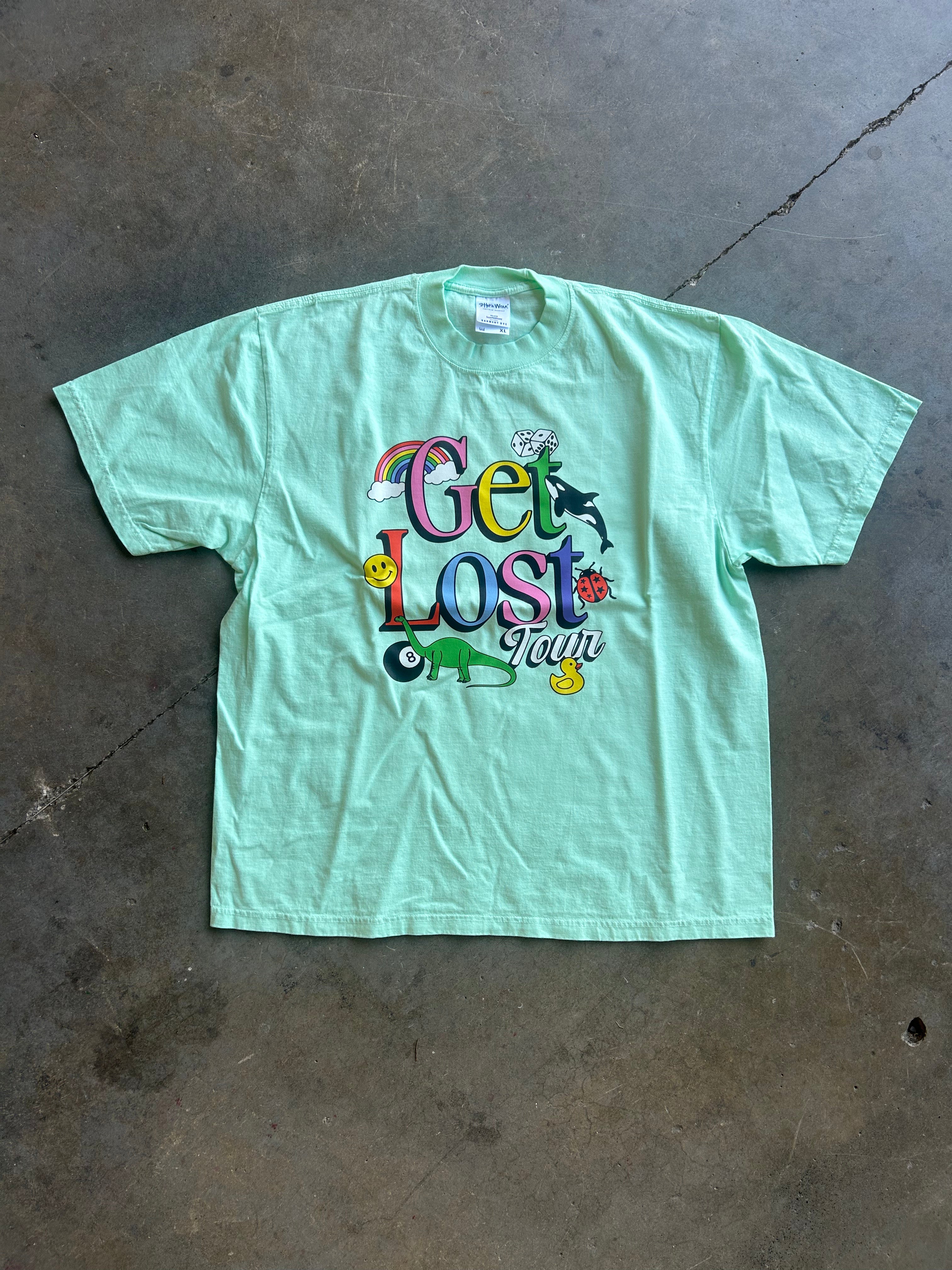 A mint green short-sleeve t-shirt with a colorful 'Get Lost Tour' graphic print, laid flat on a gray concrete floor.