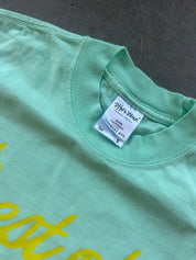 Close-up of a mint green t-shirt showing the inner neck label which reads 'Shaka Wear' and a yellow script graphic partially visible on the front.