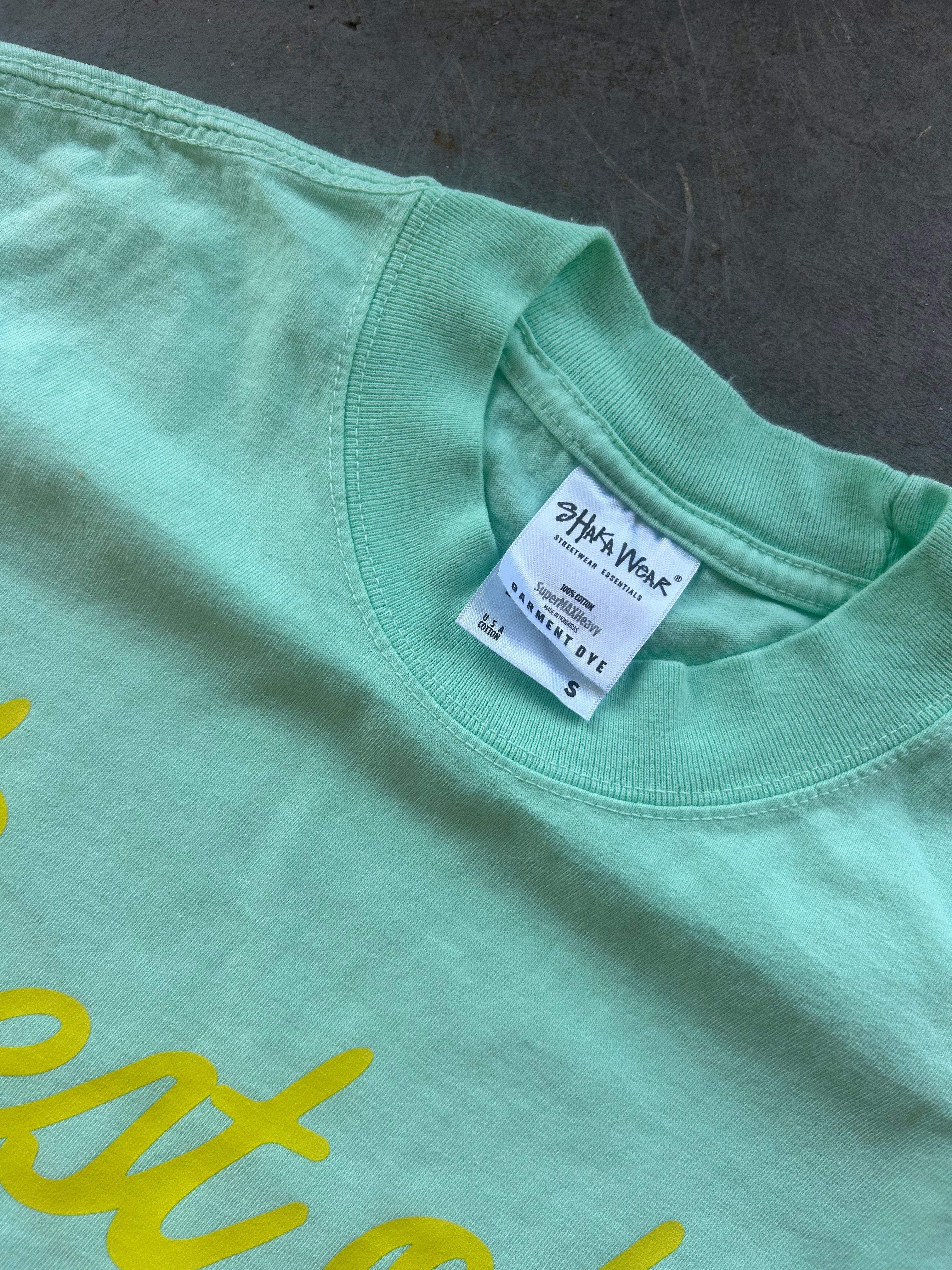 Close-up of a mint green t-shirt showing the inner neck label which reads 'Shaka Wear' and a yellow script graphic partially visible on the front.