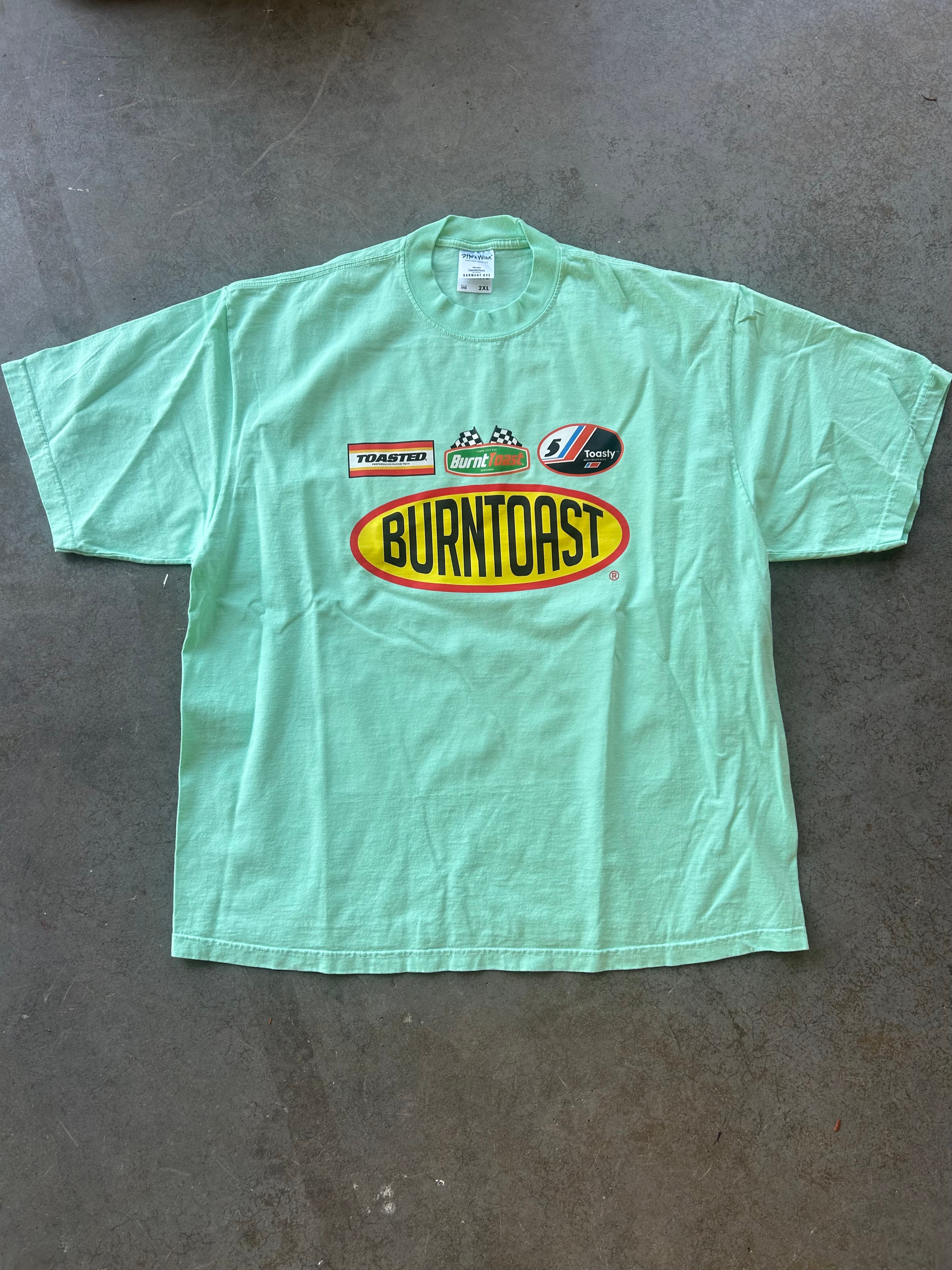 A mint green short-sleeve t-shirt laid flat on a gray concrete floor, featuring a large 'Burntast' oval logo in yellow and red on the chest, along with smaller sponsor logos above it.