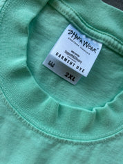 Close-up of a mint green garment dye label on a ribbed knit fabric, showing the brand 'Shaka Wear' and size '2XL'.