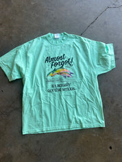 A mint green graphic t-shirt laid flat on a concrete floor. The shirt features a colorful fish illustration and the text 'Almost Forgot! it's actually not that serious.