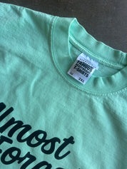 A close-up of a mint green t-shirt laid flat on a dark gray surface, showing the inner neck label which reads 'FAMOUS' and 'COTTON BLEACHED T-SHIRTS' and '2XL'.