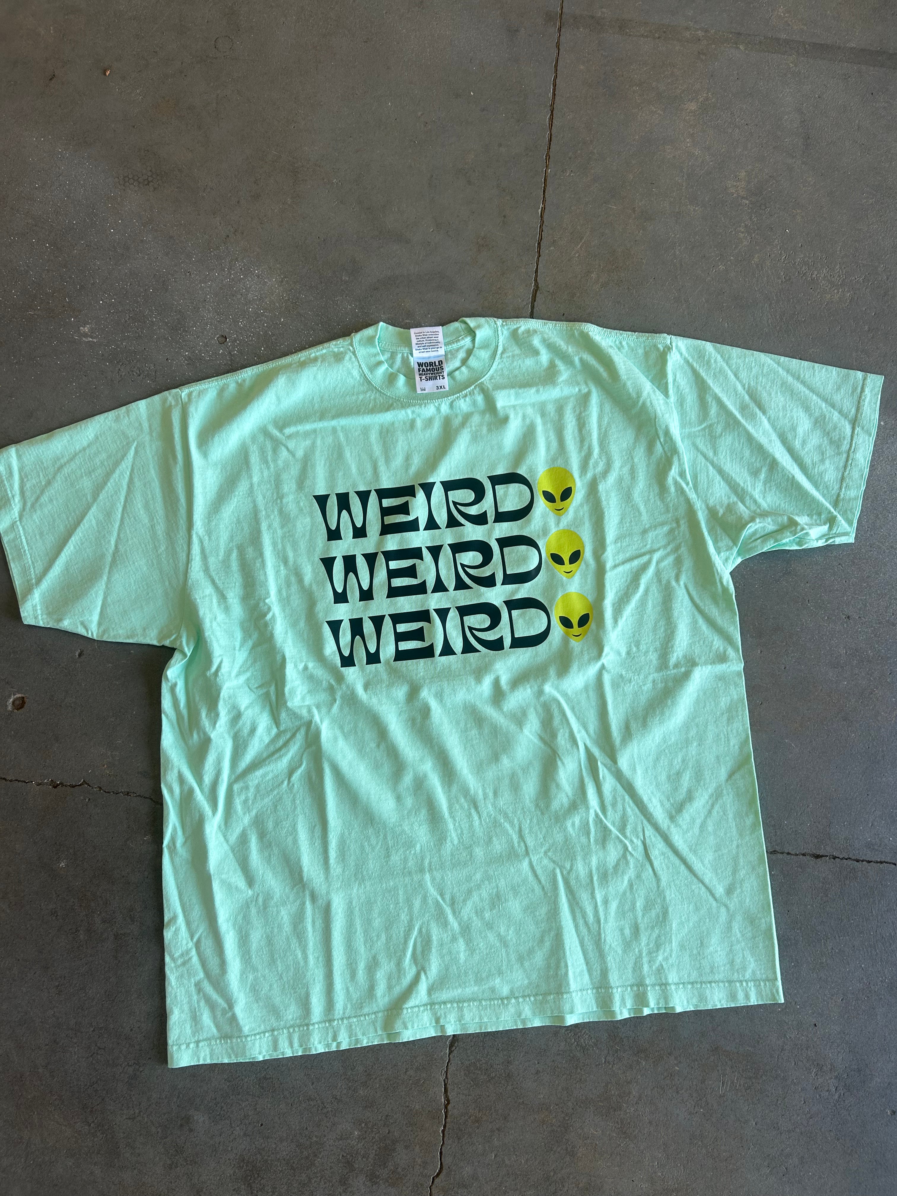 A light mint green short-sleeve t-shirt laid flat on a gray concrete floor. The front of the shirt features the word 'WEIRD' printed three times in black, with a yellow alien head graphic next to each word.