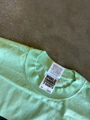 A close-up of a mint green t-shirt laid flat on a textured concrete floor, showing the inner neck label which reads 'WORLD FAMOUS EARTHWEAR T-SHIRTS'.