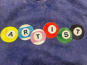 A close-up of a blue mineral-wash t-shirt featuring a graphic spelling 'ARTIST' in colorful circular letters (yellow, blue, pink, red, green, black) on the chest.