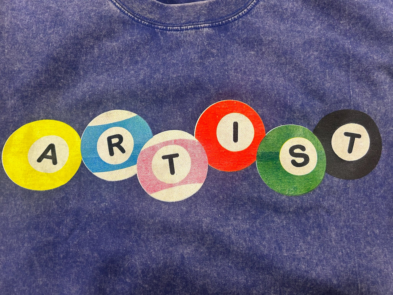 A close-up of a blue mineral-wash t-shirt featuring a graphic spelling 'ARTIST' in colorful circular letters (yellow, blue, pink, red, green, black) on the chest.