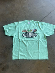 A light mint green short-sleeve t-shirt laid flat on a concrete floor. The shirt features a large oval 'BURNTOAST' logo printed on the chest, with smaller sponsor logos above it, including 'Sprintwest'.
